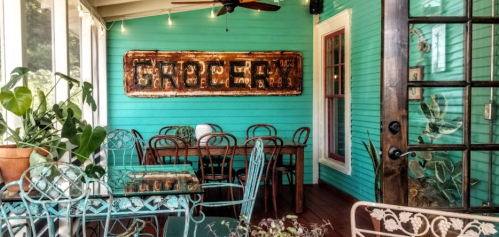 A bright turquoise room with a rustic "Grocery" sign, featuring vintage chairs and a glass table surrounded by plants.