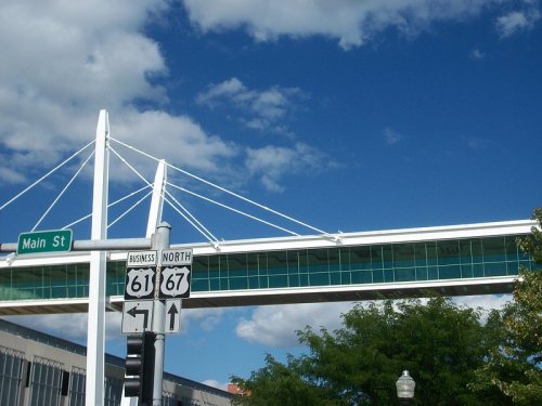 A pedestrian bridge with a modern design spans over a street sign, under a blue sky with scattered clouds.