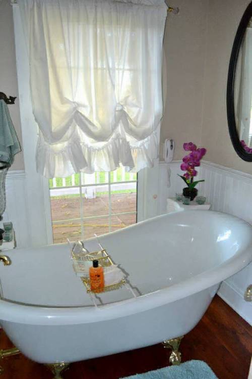 A bright bathroom featuring a white clawfoot tub, a window with sheer curtains, and a small table with toiletries.