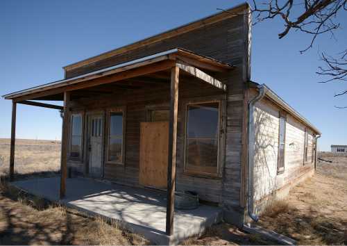 Abandoned wooden house with a porch, boarded windows, and a clear blue sky in a rural landscape.