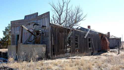 Abandoned wooden house with a collapsed roof, broken windows, and a barren landscape in the background.