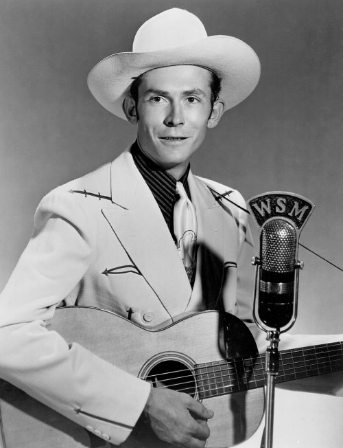 A man in a white suit and cowboy hat holds a guitar, smiling beside a vintage microphone labeled "WSM."
