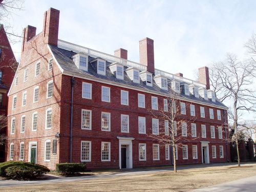 A large, historic brick building with multiple windows and chimneys, set in a grassy area under a clear sky.