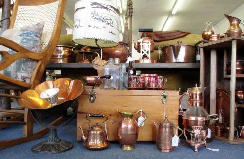 A collection of vintage copper items, including teapots, a lamp, and a wooden chest, displayed in an antique shop.
