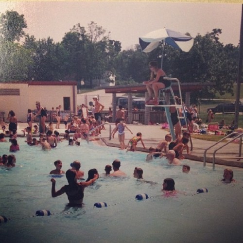 A busy outdoor pool scene with children swimming, a lifeguard on a chair, and people relaxing around the pool.