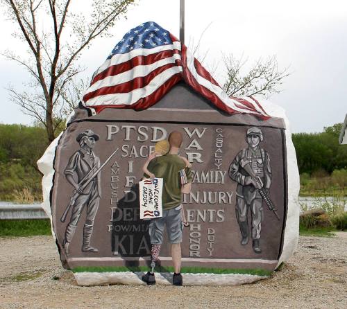 A person stands by a large rock memorial featuring military figures and inscriptions about PTSD and war casualties.