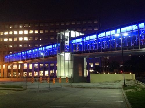 A modern building with a blue-lit skybridge connecting two structures at night, surrounded by a parking lot.