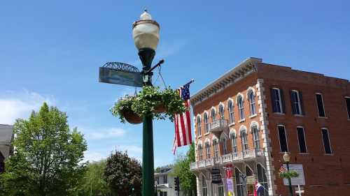 A street lamp adorned with flowers and an American flag, beside a historic brick building under a clear blue sky.