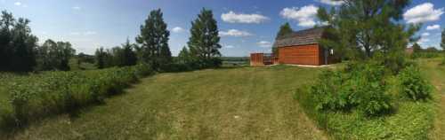 A panoramic view of a grassy field with a wooden cabin and trees under a clear blue sky.