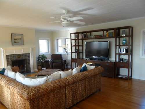 Cozy living room with a large woven sofa, a TV unit, fireplace, and a ceiling fan, featuring warm wooden floors.