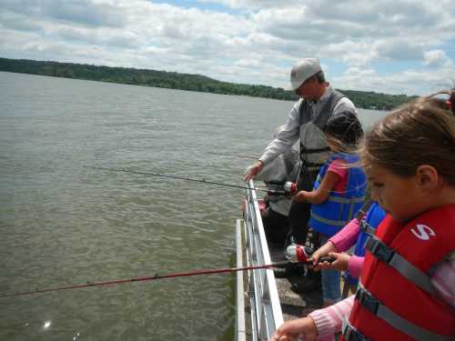 A man and two children fish from a dock, holding fishing rods over the water on a cloudy day.