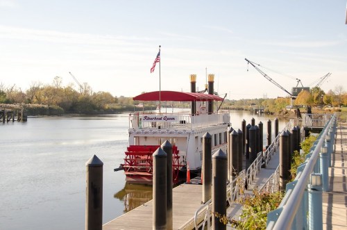A riverboat docked at a pier, with a calm river and cranes in the background under a clear blue sky.