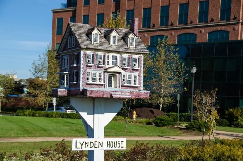 A miniature house on a post, labeled "Lynden House," set against a backdrop of buildings and greenery.