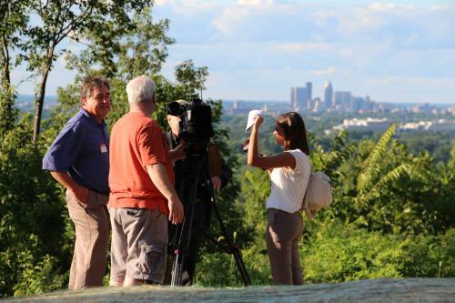 A news crew films outdoors with a city skyline in the background, featuring three people and lush greenery.