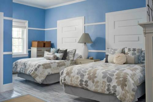 A bright blue bedroom featuring two twin beds with floral bedding, a lamp, and a dresser against the wall.