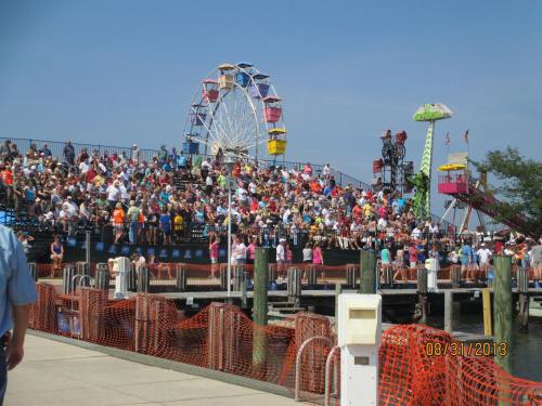 A crowded amusement park with a large ferris wheel and colorful rides, set against a clear blue sky.