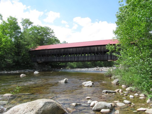 A wooden covered bridge spans a river, surrounded by lush greenery and rocky riverbanks under a blue sky.