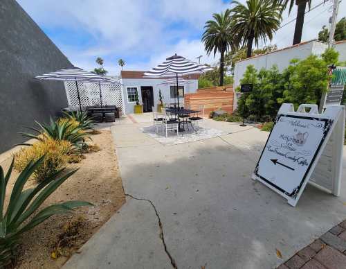 A sunny outdoor patio with tables, umbrellas, and greenery, featuring a sign pointing to a nearby entrance.