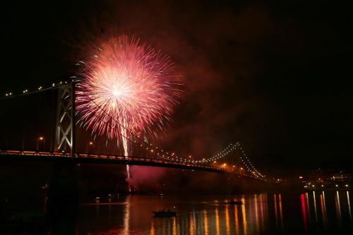 Colorful fireworks burst over a bridge at night, reflecting in the water below.