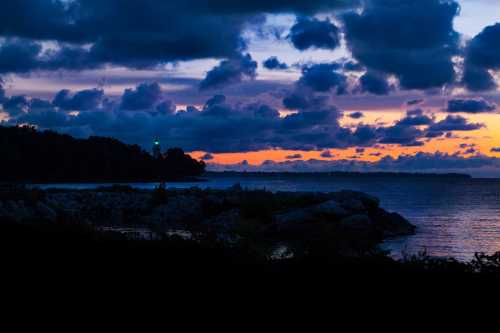 A serene sunset over a rocky shoreline, with vibrant clouds and a distant lighthouse glowing against the twilight sky.