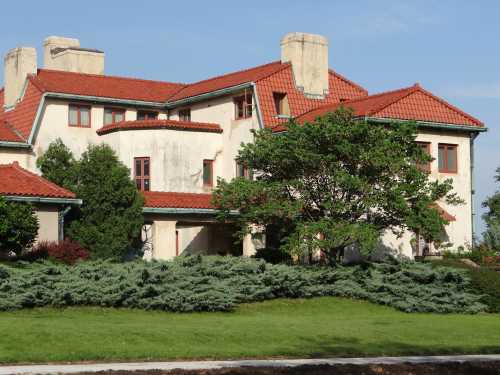 A large, two-story house with a red tile roof, surrounded by green shrubs and trees under a clear blue sky.