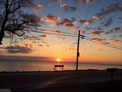 A serene sunset over a calm lake, with clouds in the sky and a bench by the roadside.