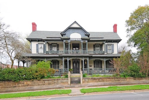 A large, historic Victorian-style house with a wraparound porch and decorative trim, surrounded by greenery.