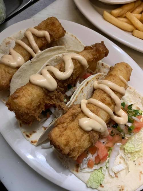 Three crispy fish tacos topped with sauce, served on a plate with fresh vegetables and a side of fries.