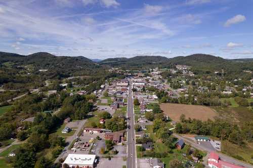 Aerial view of a small town surrounded by green hills and fields, with a main road running through the center.