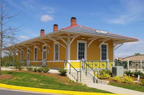Historic yellow train station with a red roof, surrounded by greenery and a clear blue sky.