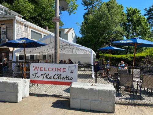 A patio area with tables and umbrellas, featuring a sign that says "Welcome To The Chatio." Trees in the background.