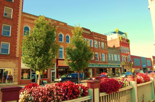 A charming street scene featuring brick buildings, green trees, and colorful flower beds in a small town.