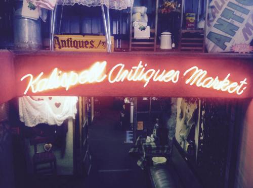 Neon sign reading "Kalispell Antiques Market" above an entrance, surrounded by vintage decor and antiques.