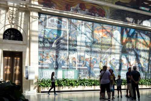 A group of people gathers in front of a large mural depicting industrial scenes, with plants and sunlight filtering in.