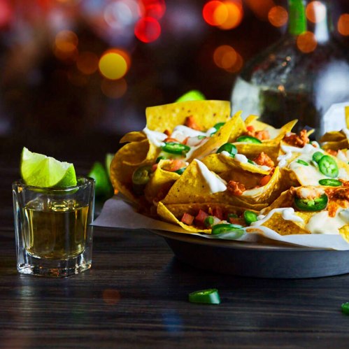 A plate of nachos topped with jalapeños and sour cream, alongside a shot of tequila and a lime wedge.