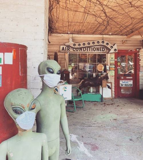 Two green alien figures wearing masks stand outside a quirky, rustic building with an "Air Conditioned" sign.