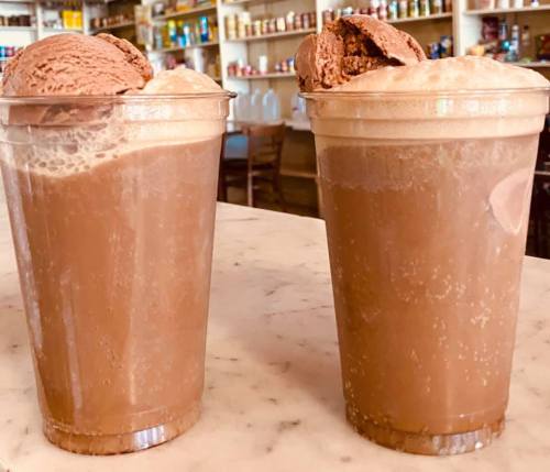 Two clear cups filled with chocolate milkshake topped with scoops of chocolate ice cream, sitting on a marble counter.