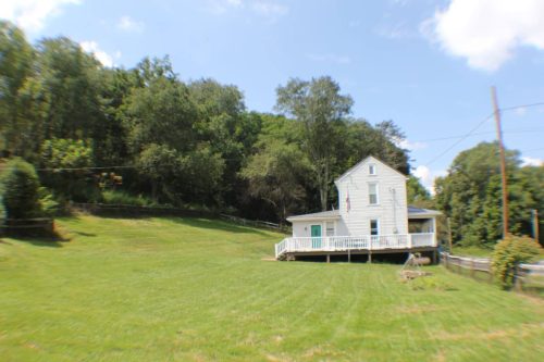 A white two-story house on a grassy hill, surrounded by trees under a blue sky with fluffy clouds.