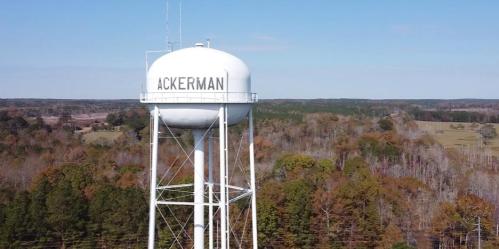 A white water tower with "ACKERMAN" written on it, surrounded by trees and a clear blue sky.