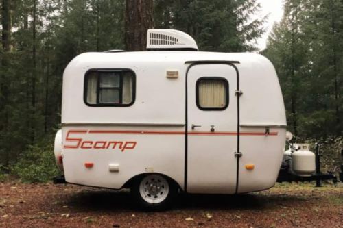 A small white Scamp trailer parked in a forest, surrounded by tall trees and a gravel ground.