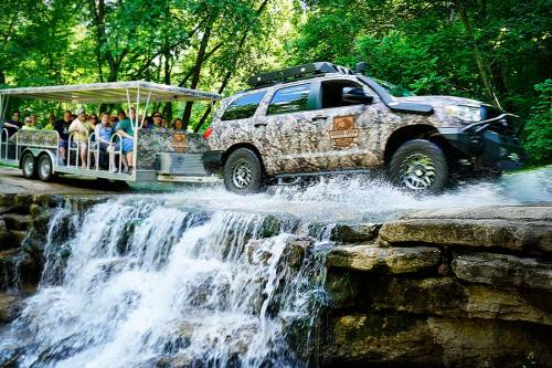 A camouflaged vehicle pulls a trailer with passengers over a small waterfall in a lush, green forest.