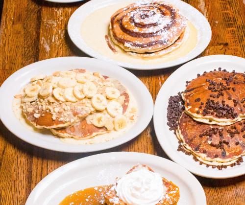 A variety of pancakes topped with bananas, chocolate chips, and cinnamon, served on white plates on a wooden table.