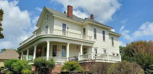 A large, two-story white house with a wraparound porch and green landscaping under a partly cloudy sky.