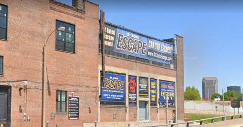 Exterior of a brick building featuring signs for an escape room business, with a city skyline in the background.