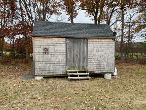 A small wooden shed with a sloped roof, steps, and a weathered exterior, surrounded by autumn foliage.