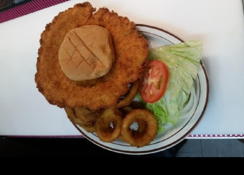 A plate featuring a large breaded patty topped with a bun, accompanied by lettuce, tomato, and onion rings.