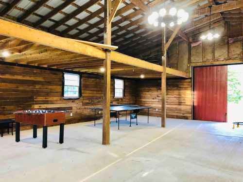 Interior of a rustic barn with wooden walls, featuring a foosball table and a large table under bright lights.