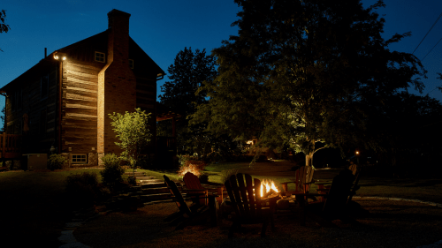 A cozy outdoor scene at night featuring a fire pit surrounded by chairs, with a house and trees in the background.