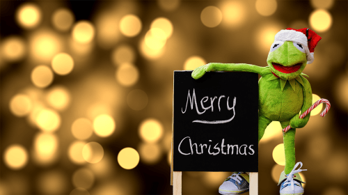 Kermit the Frog in a Santa hat holds a sign that says "Merry Christmas," with a festive bokeh background.