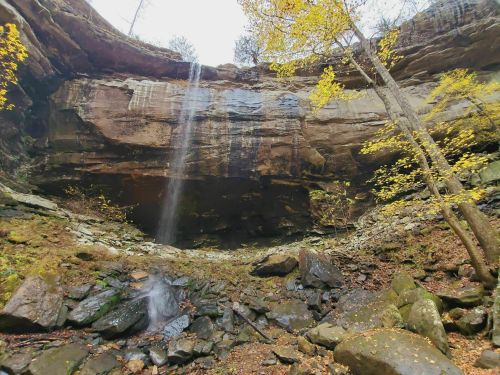 A serene waterfall cascades from a rocky overhang, surrounded by autumn foliage and scattered boulders.
